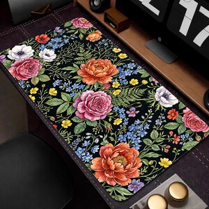 Puede incluir: Alfombrilla de escritorio rectangular negra con un vibrante estampado floral. El dise&ntilde;o presenta grandes flores coloridas en tonos rosa, naranja y blanco, junto con acentos azules y amarillos. Hojas y tallos verdes completan el tema bot&aacute;nico.