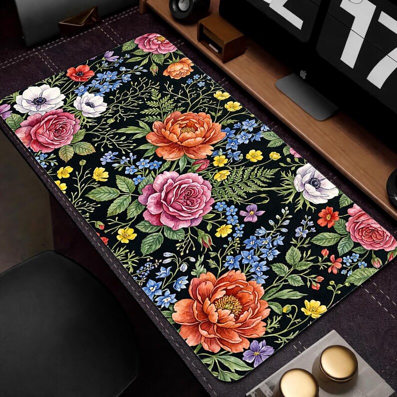 Puede incluir: Alfombrilla de escritorio rectangular negra con un vibrante estampado floral. El dise&ntilde;o presenta grandes flores coloridas en tonos rosa, naranja y blanco, junto con acentos azules y amarillos. Hojas y tallos verdes completan el tema bot&aacute;nico.