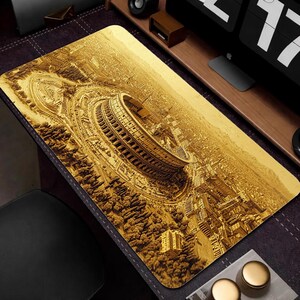 May include: A rectangular desk mat featuring a detailed, sepia-toned aerial view of the Colosseum in Rome. The design includes surrounding buildings, roads, and trees, creating a textured, three-dimensional effect. The mat is placed on a dark surface.