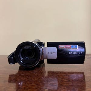 Pode incluir: Uma filmadora Samsung preta com lente Schneider Kreuznach, porta HDMI e painel lateral com etiquetas para reprodução Full HD 1080, zoom de 65x a 100x e OIS. O dispositivo possui uma dobradiça prateada e uma alça preta na lente.