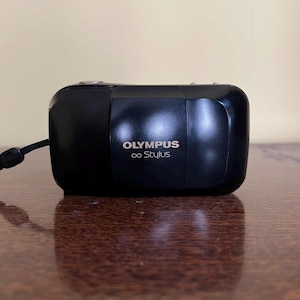 May include: A black Olympus ∞ Stylus camera with a wrist strap. The camera is rectangular with rounded edges and the brand name and model are printed in white on the front. The camera is sitting on a brown surface.