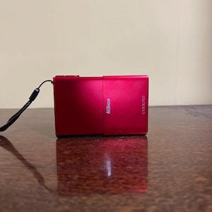 May include: A red Nikon Coolpix digital camera with a black wrist strap. The camera is rectangular with the Nikon and Coolpix brand names printed on the side. The camera is on a wooden surface.