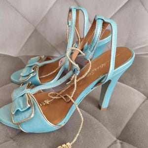 May include: A pair of light blue high-heeled sandals featuring a prominent bow on the front strap, gold-toned trim, and thin gold-colored laces that wrap around the ankle. The interior sole is tan and displays the brand name Miss Sis in script.