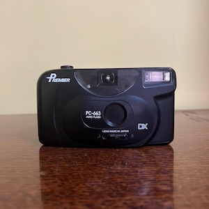 May include: A black Premier PC-663 35mm film camera with an auto flash feature. The front of the camera displays the brand name Premier, the model number PC-663, the text AUTO FLASH, the letters DX, and the text LENS MADE IN JAPAN.