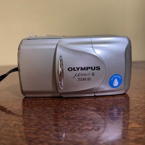 May include: Silver Olympus µ [mju:]-II Zoom 80 camera with a black strap. The camera has the text "OLYMPUS" and "ALL WEATHER" printed on it. The camera is on a reflective brown surface.