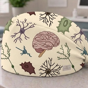Può includere: Una cuffia chirurgica color crema con un motivo ripetuto di cervelli anatomici, vari tipi di neuroni e strutture cellulari in tonalità rosa, verde, viola e marrone. La cuffia presenta una chiusura a laccetti e una piccola etichetta bianca.