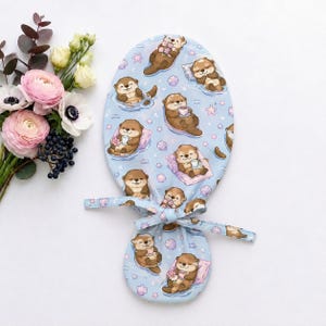 May include: A light blue fabric scrub cap featuring a repeating pattern of brown otters, pink seashells, and small stars. The otters are depicted in various poses, some holding pink flowers or white cups, and others resting on small pink pillows.