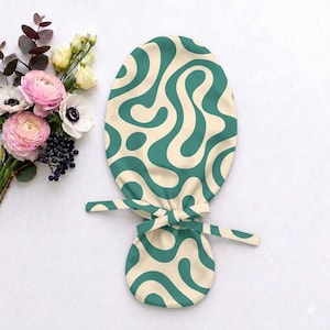 May include: A scrub cap featuring a wavy, abstract pattern in teal and cream. The cap has a rounded top section and a smaller, circular lower section connected by fabric ties that are knotted in the centre to secure the fit around the head.