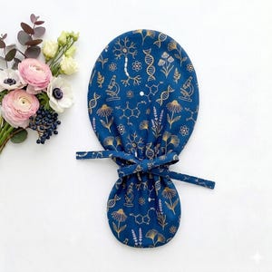 Op de afbeelding: Een blauwe operatiemuts met een goudkleurig patroon van wetenschappelijke illustraties, waaronder DNA-helices, chemische structuren, neuronen, microscopen en diverse botanische elementen zoals bloemen en bladeren, vastgebonden met stoffen linten.