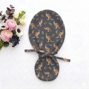 May include: A dark gray scrub cap featuring a repeating pattern of brown kangaroos and small yellow floral sprigs. The cap has a rounded top section and a smaller lower section, connected by two fabric ties that are knotted in the center.