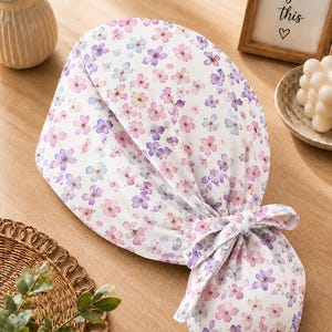Può includere: Una cuffia chirurgica bianca con un delicato motivo floreale composto da piccoli fiori rosa e viola. La cuffia presenta una parte superiore arrotondata e una sezione posteriore arricciata, fissata con un laccetto in tessuto.