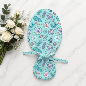 May include: A light blue scrub cap featuring a repeating pattern of colorful cells, bacteria, and microscopic organisms in shades of purple, pink, and teal, with fabric ties at the back for an adjustable fit and a rounded top design.
