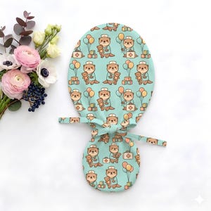May include: A light teal scrub cap featuring a repeating pattern of cartoon otters dressed as nurses, holding teddy bears and medical bags, accompanied by colorful balloons and alphabet blocks, with fabric ties for a secure fit.