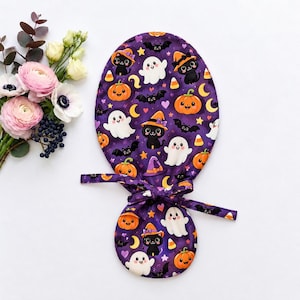 May include: A purple fabric scrub cap featuring a repeating pattern of black cats wearing orange witch hats, white ghosts, orange pumpkins, black bats, yellow stars, purple hearts, and candy corn. The cap has a tie-back closure with fabric straps.