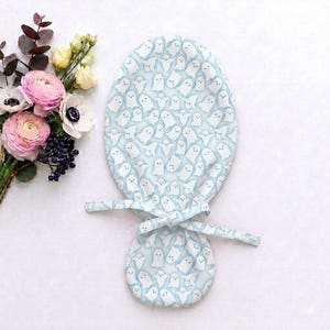 May include: A light blue fabric scrub cap featuring an all-over pattern of small white ghosts. The cap has a rounded top section and a smaller circular back section, connected by fabric ties that are knotted in the center to secure the fit.