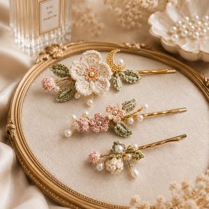 May include: Three gold-toned metal hair clips featuring crocheted floral designs in cream, soft pink, and sage green. Each clip is embellished with small white faux pearls and one clip includes a small gold-toned crescent moon charm.