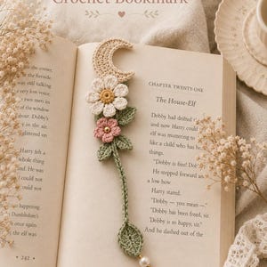 Può includere: Un segnalibro all'uncinetto con una mezzaluna beige, una margherita bianca con centro giallo, un piccolo fiore rosa con una perlina dorata e foglie verdi su uno stelo. Il segnalibro termina con una foglia verde e una perlina bianca.