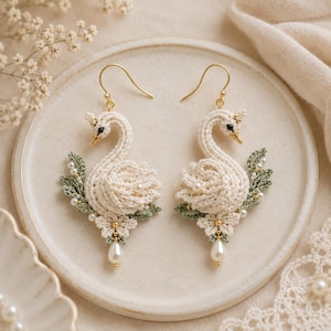 Pode incluir: Um par de brincos pendentes com cisnes de crochê na cor creme, olhos pretos e bicos dourados. Cada cisne é decorado com folhas de crochê verde sálvia, pequenas flores brancas, minúsculas pérolas e um pingente de pérola em forma de gota.