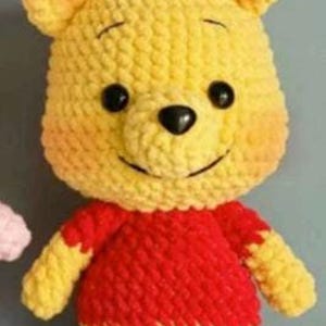 Puede incluir: Un muñeco de Winnie the Pooh hecho a mano. El oso es principalmente amarillo con una camisa roja y ojos negros. El muñeco tiene una expresión sonriente y está hecho de un hilo suave y texturizado.