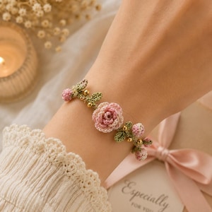 May include: A crochet bracelet featuring a central pink rose, smaller pink flower buds, and green leaves, accented with small gold-toned beads. The floral design is crafted from fine thread and is secured around the wrist with a delicate, flexible band.