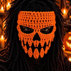 Peut inclure: Une cagoule en crochet noir présentant un motif de crâne orange avec des découpes pour les yeux et le nez. Le masque comprend de longues franges noires en forme de corde sur le dessus et les côtés, ressemblant à des cheveux, couvrant la tête.