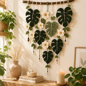 May include: A wall hanging featuring crocheted dark green monstera leaves and white flowers with yellow centres, suspended from a horizontal wooden branch by thin twine. The arrangement includes small green leaves and beads along the hanging cords.