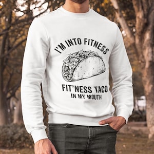 Può includere: Felpa bianca con la scritta "I'M INTO FITNESS" sopra una grafica di taco e "FIT'NESS TACO IN MY MOUTH" sotto. La felpa a maniche lunghe è indossata con jeans scuri.