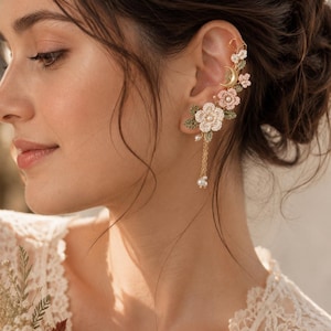 May include: An ear cuff featuring delicate crochet flowers in shades of white and soft pink, accented with green crocheted leaves, a small gold-toned crescent moon charm, and dangling gold chains finished with tiny white pearl-like beads.
