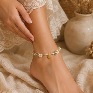 May include: An anklet featuring a delicate chain of small white pearls interspersed with tiny crocheted white flowers, green leaves, and a central gold-toned crescent moon charm with an intricate filigree pattern, secured with a simple clasp.