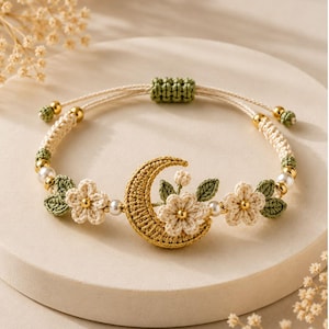 May include: A macrame bracelet featuring a central gold-coloured crescent moon charm adorned with cream-coloured crocheted flowers and green leaves. The band includes small white pearls, gold-toned beads, and an adjustable green sliding knot closure.