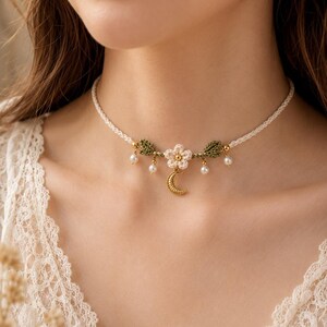 May include: A cream-colored braided choker necklace featuring a central crocheted flower, two green crocheted leaves, and a small gold-toned crescent moon charm. The design is accented with four small white spherical beads hanging from gold-toned links.