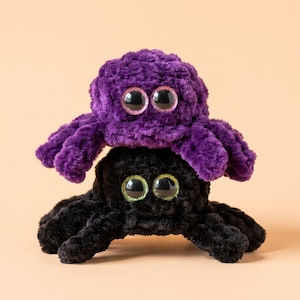 Puede incluir: Dos arañas de peluche, una morada y otra negra, están apiladas. La araña morada está encima. Ambas tienen ojos grandes y brillantes. Los juguetes están hechos de un material suave y texturizado, sobre un fondo melocotón claro.