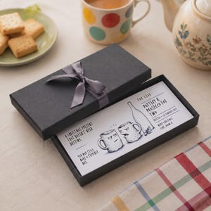 May include: A black rectangular gift box with a grey ribbon, containing a white card with a cartoon illustration of two mugs and a bottle. The text reads: For Leah, Pottery & Prosecco for two, Bristol 25 January 2027, Merry Christmas Love, Naomi x.