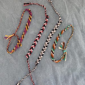May include: Three handmade woven bracelets featuring intricate patterns. One bracelet displays purple, yellow, and orange diagonal stripes. Another features a black, white, and red chevron design. The third shows green, brown, and white colors.