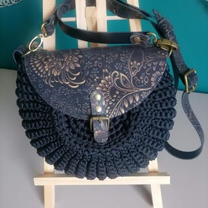 Può includere: Borsa a tracolla all'uncinetto blu navy con chiusura a patta. La patta presenta un motivo floreale in oro e blu navy. La borsa ha una tracolla regolabile abbinata e una chiusura con fibbia. La borsa è esposta su un piccolo cavalletto di legno.