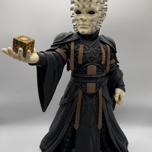 May include: A pale-skinned figurine with numerous silver pins protruding from the head, wearing a black robe with brown accents. The figure holds a small, square, gold and black patterned puzzle box in an outstretched hand.