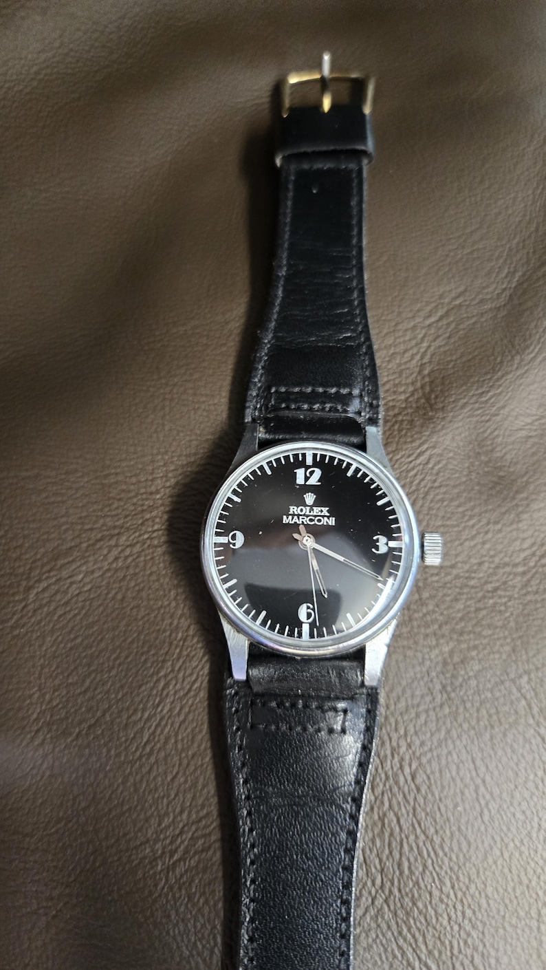 May include: A black-faced Rolex Marconi wristwatch with a silver case and a black leather band. The watch face features white numerals at the 12, 9, and 6 o'clock positions. The watch band has a gold-colored buckle.