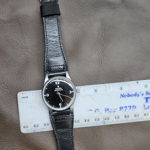May include: A black-faced Rolex Marconi wristwatch with a black leather band. The watch has silver hands and hour markers. The watch is measured with a ruler. The watch band is approximately 1 inch wide.