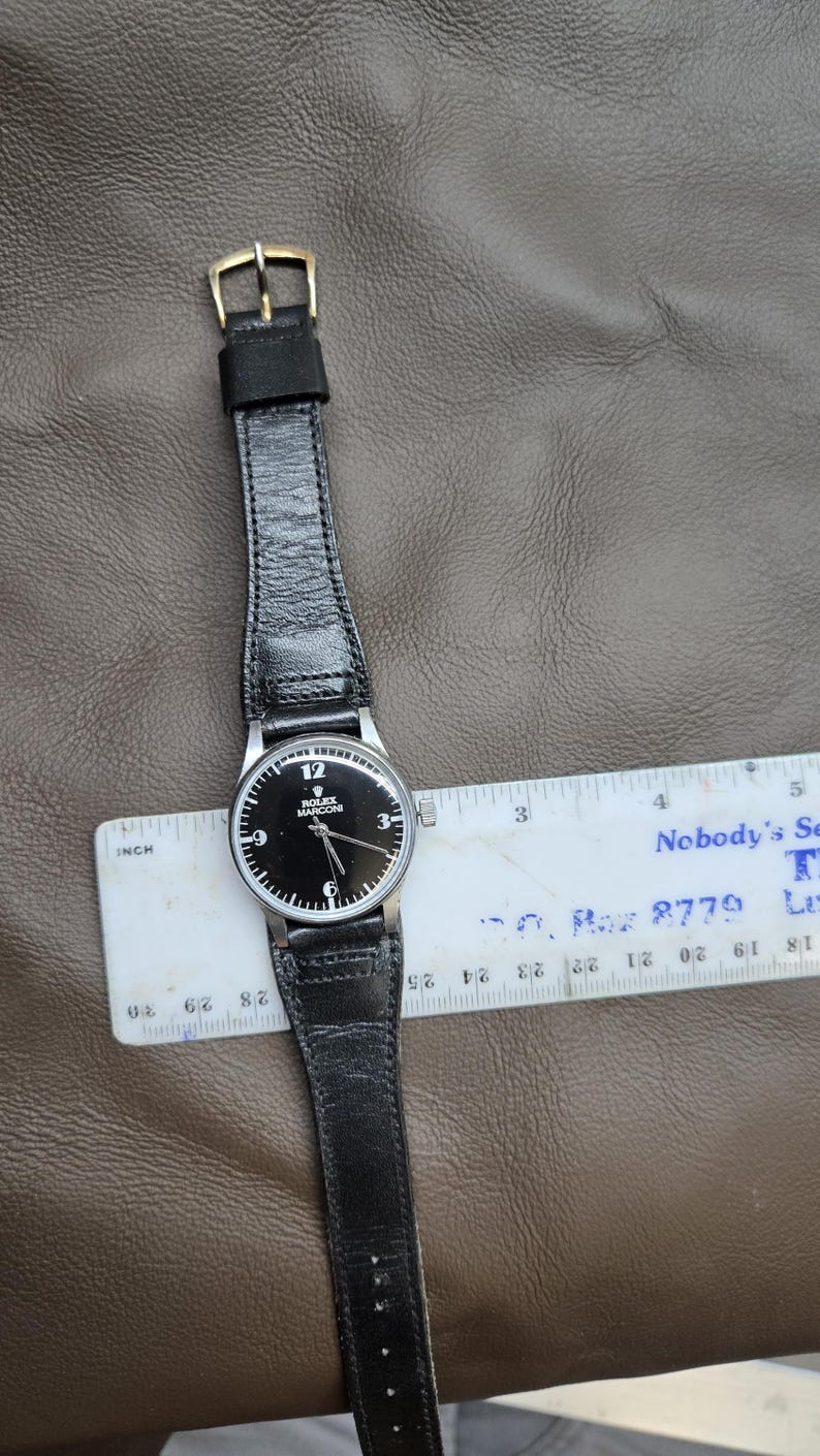 May include: A black-faced Rolex Marconi wristwatch with a black leather band. The watch has silver hands and hour markers. The watch is measured with a ruler. The watch band is approximately 1 inch wide.