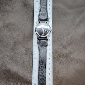 May include: A black wristwatch with a black leather band, silver case, and a black watch face. The watch is measured with a ruler, showing the band is approximately 10 inches long.