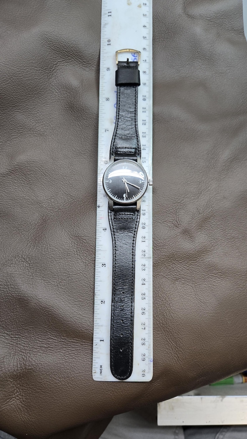May include: A black wristwatch with a black leather band, silver case, and a black watch face. The watch is measured with a ruler, showing the band is approximately 10 inches long.