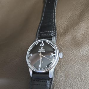 May include: A vintage Rolex Marconi wristwatch with a black dial and silver hands. The watch has a black leather strap and a silver case. The dial features the number 12, 3, 6, and 9. The watch is a classic timepiece.