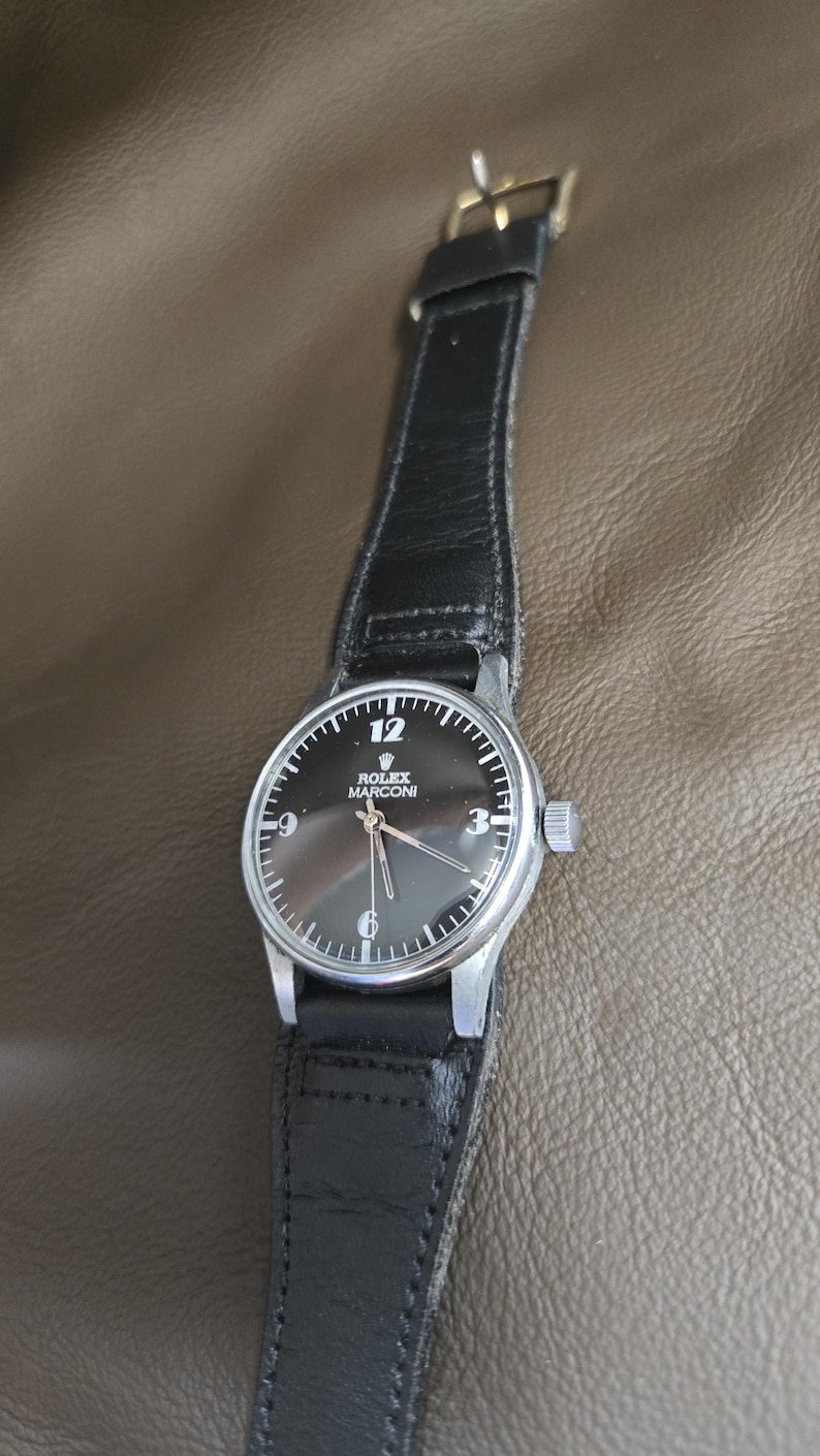 May include: A vintage Rolex Marconi wristwatch with a black dial and silver hands. The watch has a black leather strap and a silver case. The dial features the number 12, 3, 6, and 9. The watch is a classic timepiece.