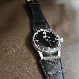 May include: A wristwatch with a black face and a silver-colored case. The watch has a black leather band. The watch face displays the number 12, the Rolex Marconi logo, and the numbers 3, 6, and 9.