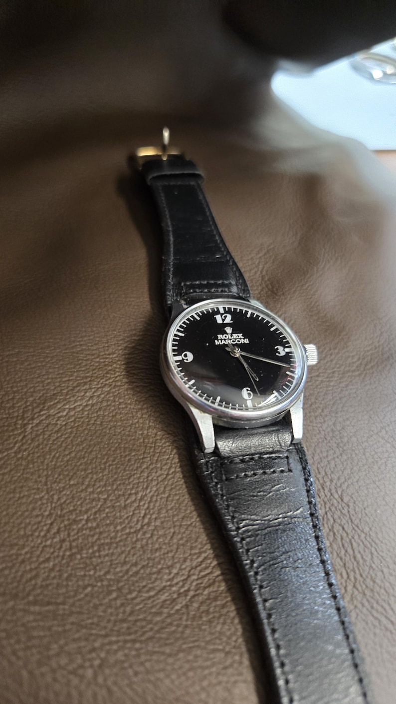 May include: A wristwatch with a black face and a silver-colored case. The watch has a black leather band. The watch face displays the number 12, the Rolex Marconi logo, and the numbers 3, 6, and 9.