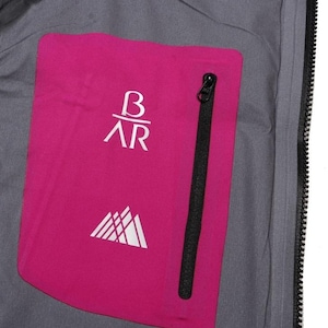 May include: Close-up of a gray jacket interior with a bright pink pocket. The pocket features white text and a mountain graphic. A black zipper runs vertically along the pocket's edge. The jacket's interior is visible.