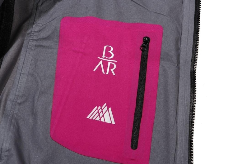 May include: Close-up of a gray jacket interior with a bright pink pocket. The pocket features white text and a mountain graphic. A black zipper runs vertically along the pocket's edge. The jacket's interior is visible.