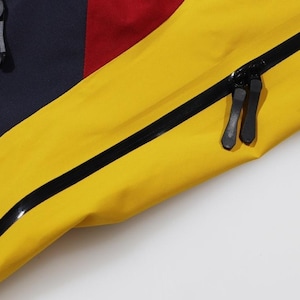 May include: Close-up of a section of a colorful jacket. The jacket features panels of navy blue, red, and bright yellow. Black zippers and trim are visible, adding contrast to the vibrant colors. The image highlights the details of the garment's construction.