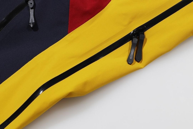 May include: Close-up of a section of a colorful jacket. The jacket features panels of navy blue, red, and bright yellow. Black zippers and trim are visible, adding contrast to the vibrant colors. The image highlights the details of the garment's construction.