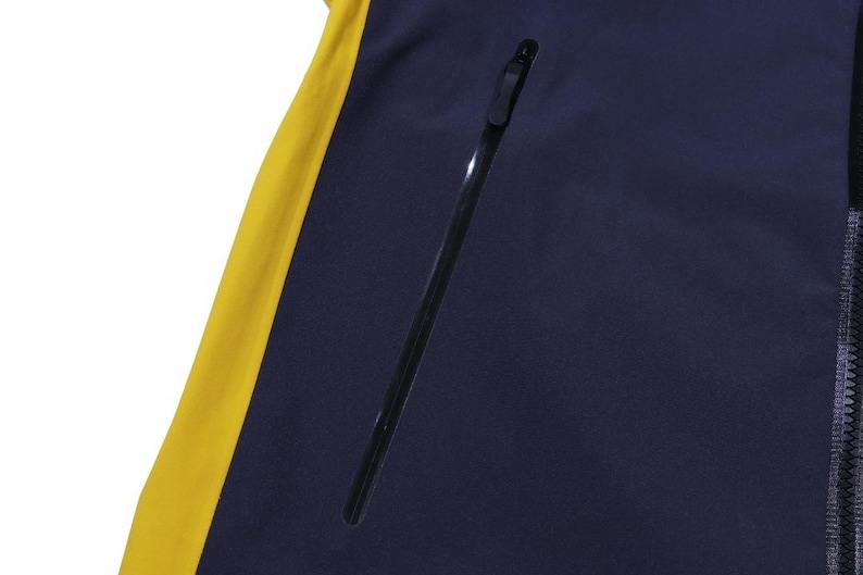 May include: Close-up of a navy blue jacket with a yellow side panel. The jacket has a black zipper and a black zippered pocket. The jacket is made of a smooth, possibly waterproof material.